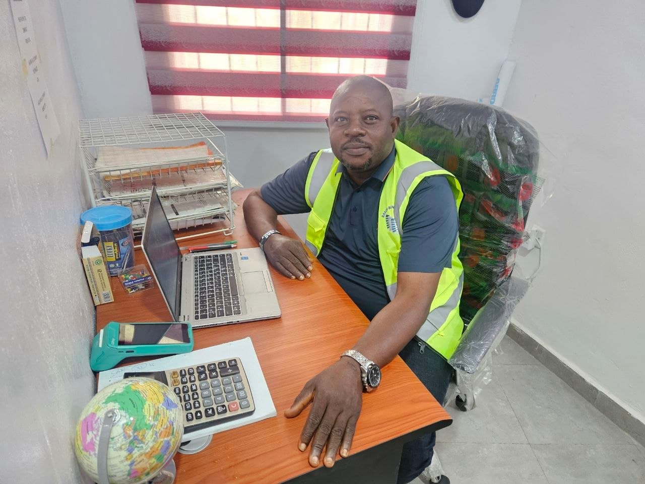 The founder of Bosun Blinds at his desk in the company office in Ibadan, wearing a branded vest, one of the company's day and night blinds fitted behind him