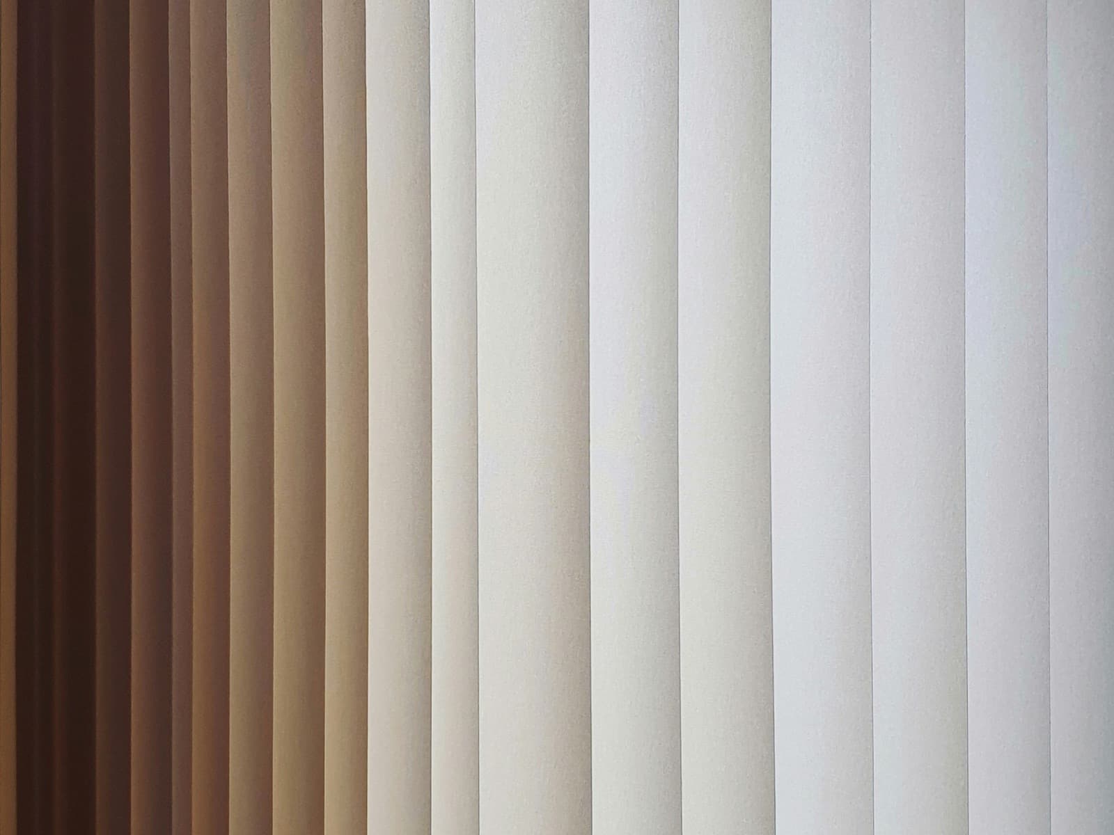 Cream vertical blind louvres angled against a window, daylight grading from warm shadow to bright across the slats